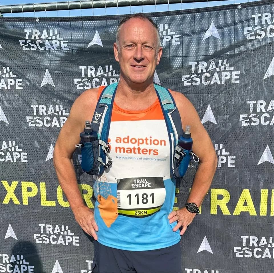 A man wearing an orange and white running vest with "adoption matters" and a race bib number 1181, posing in front of a banner that says "TRAIL ESCAPE."