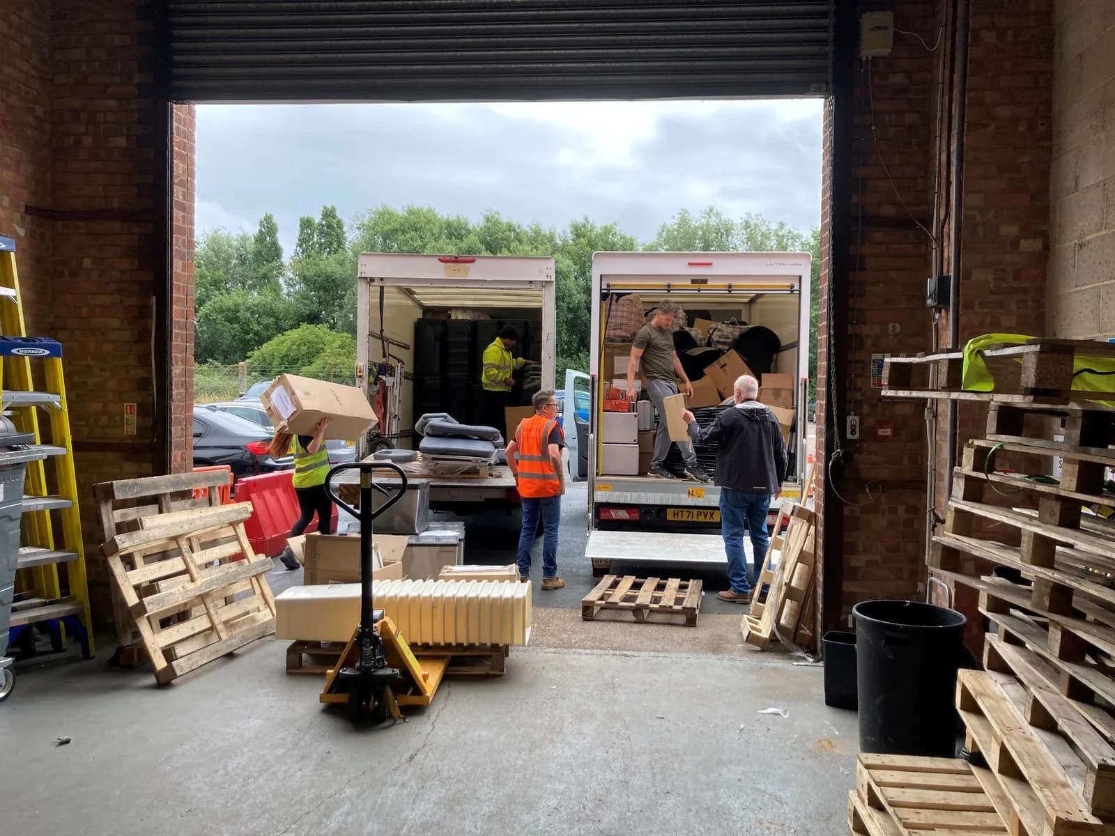 A group of people are loading boxes and other items into the back of two white delivery trucks from a warehouse loading bay.