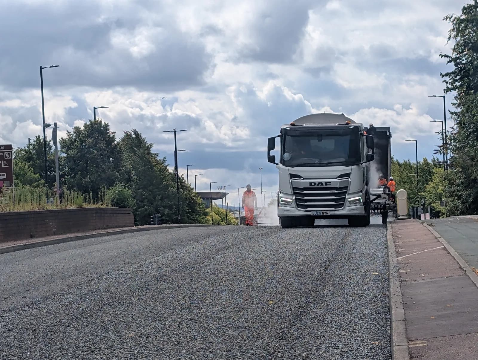 A large white DAF truck is shown paving a road, with two construction workers in orange vests nearby, and a cloudy sky above.