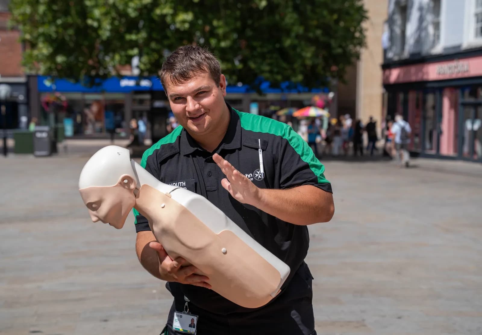 A man in a black and green uniform smiles at the camera while holding a CPR mannequin in a public square.