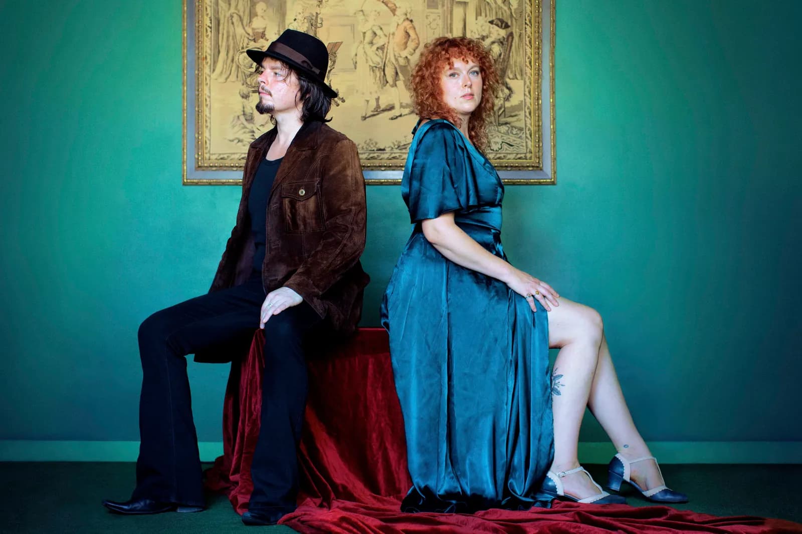 A man in a brown jacket and hat and a woman in a teal dress sitting back-to-back with a tapestry behind them.