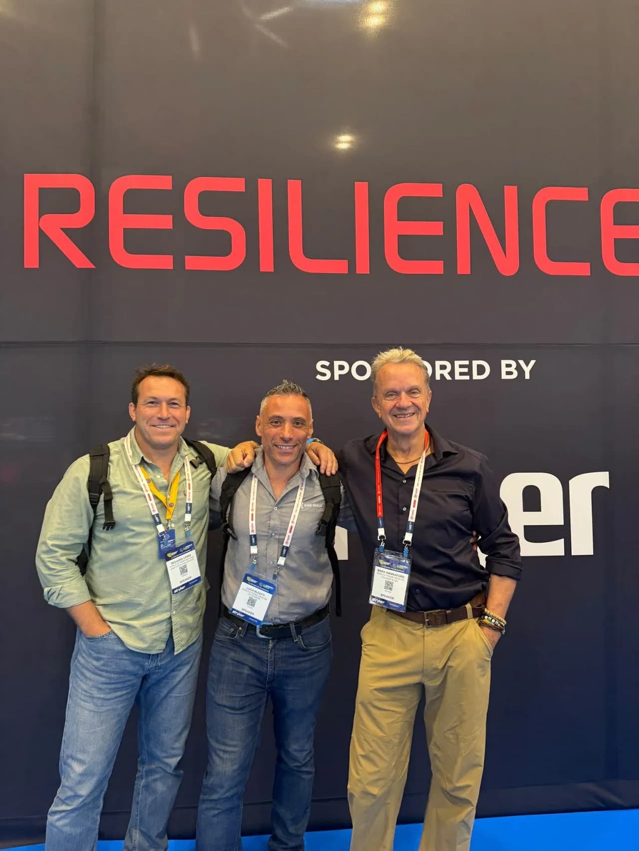 Three men stand smiling in front of a dark blue backdrop with the word "RESILIENCE" in red and other text in white.