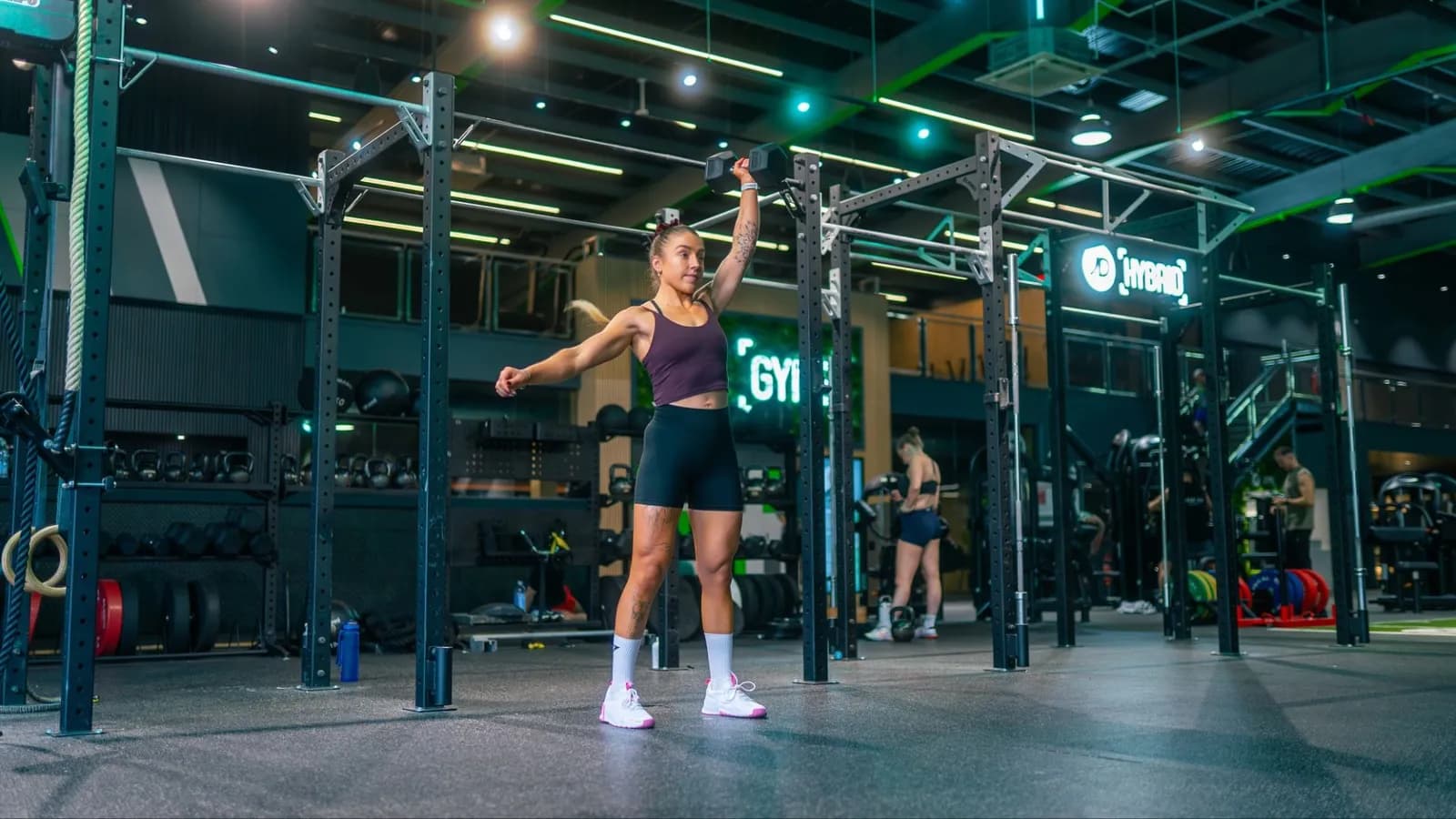 A woman in a gym performs an overhead dumbbell press with her right arm.